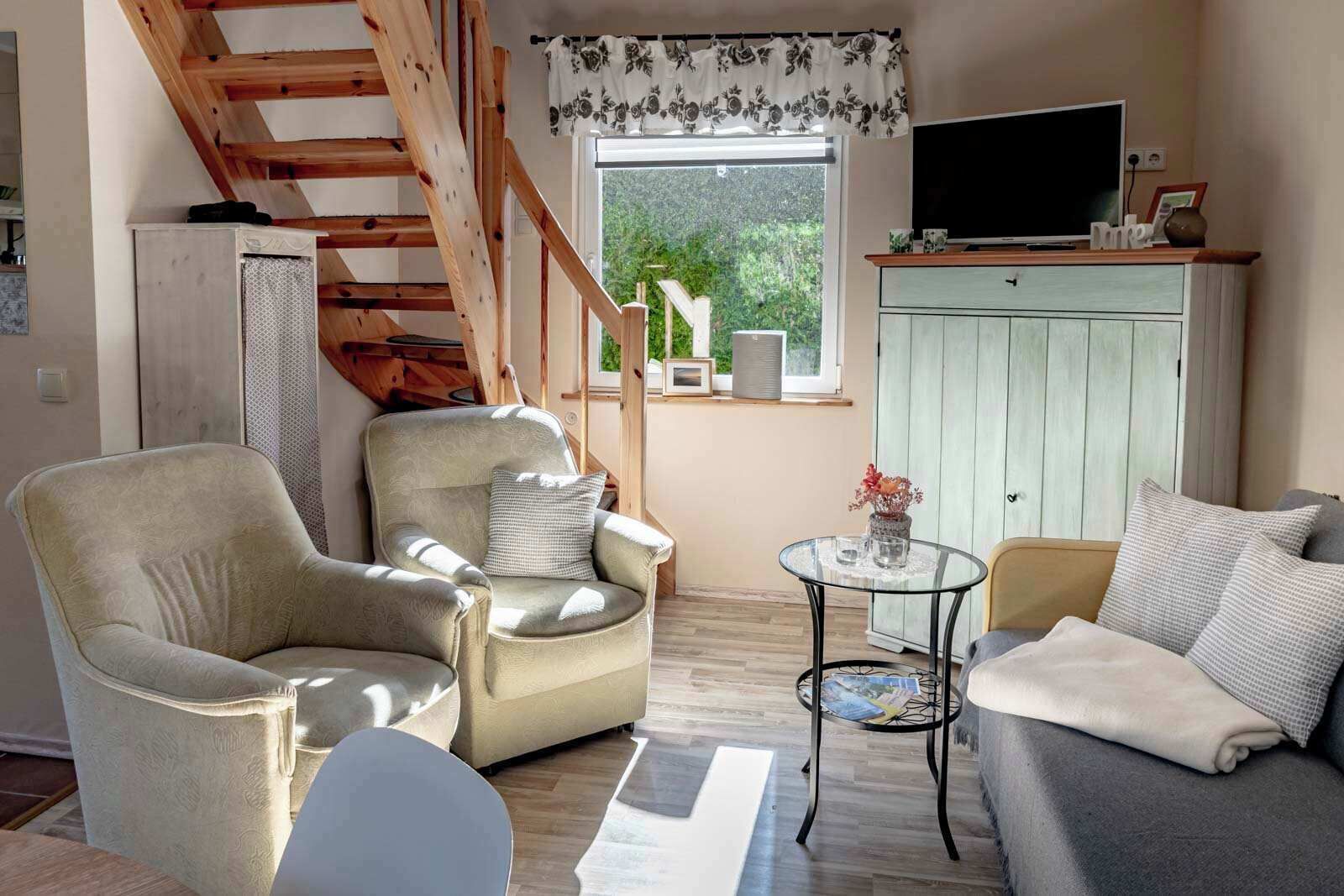 A cozy living room with a sofa, armchair, and a small table. A TV is mounted on the wall, and a staircase is visible in the background.