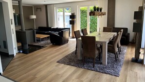 A modern living room with a black leather sofa, a wooden dining table with chairs, and a large window with a view of greenery. - Exklusives Ferienhaus Celeste nahe Warnemünde – Luxus & Entspannung für bis zu 7 Personen (Rostock)