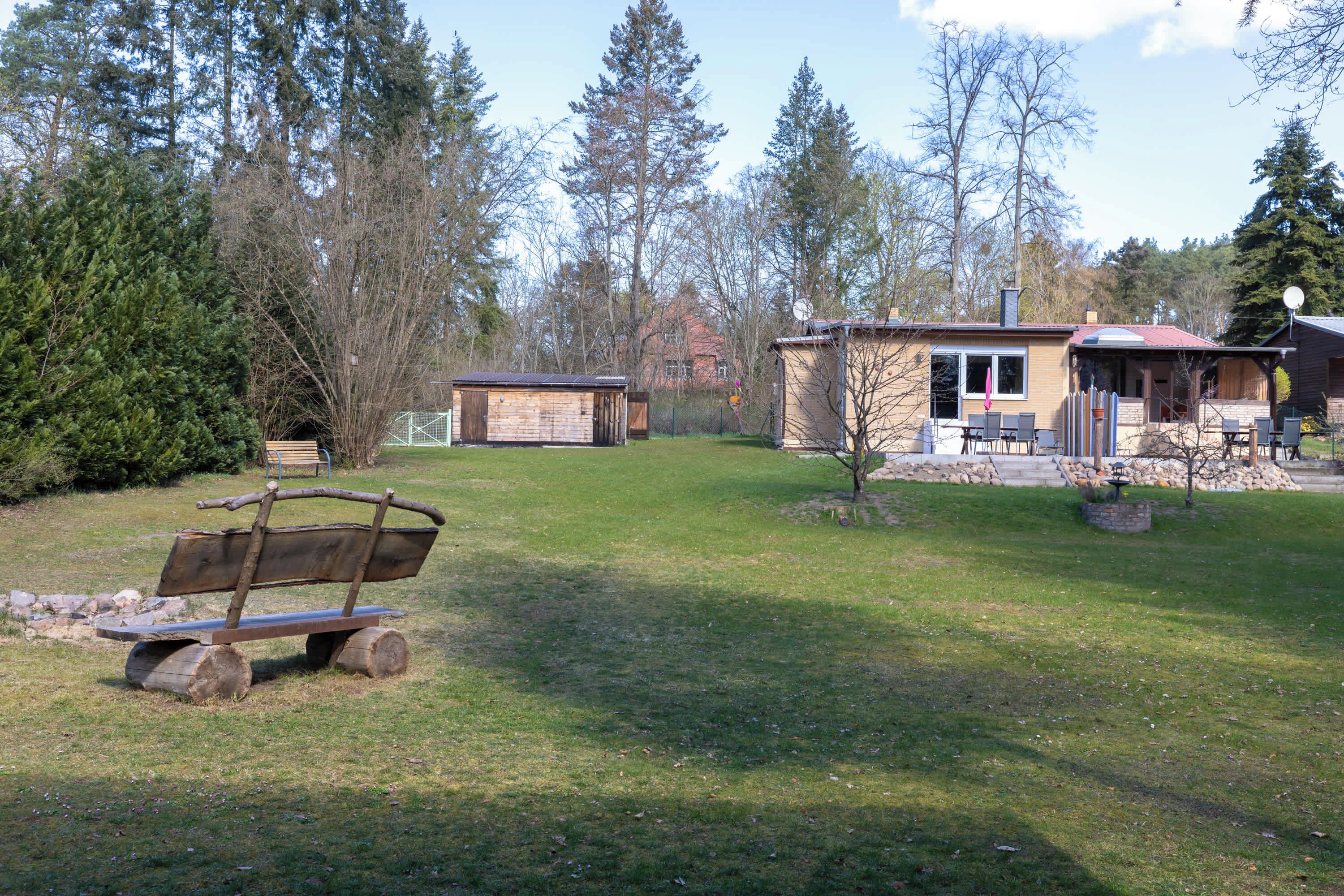 A rustic cabin with a wooden deck, surrounded by a grassy yard and tall trees.