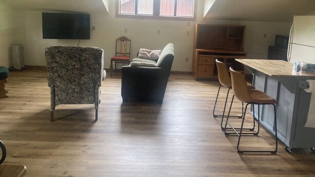 A spacious living area with a high ceiling, a large window, a flat-screen TV, a wooden dining table, and a kitchen area.