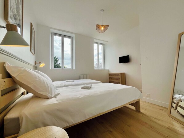 Un dormitorio con cama, mesita de noche, dos ventanas, un televisor montado en la pared y una lámpara colgante.