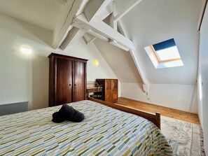 Une chambre avec un plafond en pente, une lucarne, une armoire en bois, un lit recouvert d’une couvre-lit à rayures et un banc en bois.