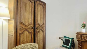Una habitación con un armario de madera, un sillón estampado, una mesita con un florero y una lámpara.