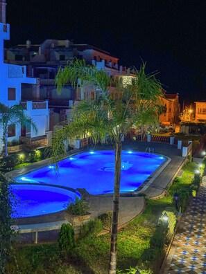Vue nocturne d’un espace piscine comprenant deux piscines, entouré de palmiers et bordé d’une allée dallée.