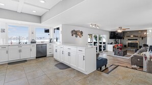 Una cocina amplia con gabinetes blancos, lavavajillas y una isla grande. Hay una zona de comedor con un sofá gris y una mesa de centro. La cocina tiene un ventanal con vistas a la ciudad.