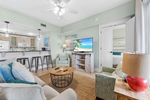 A modern living room with a sofa, coffee table, and TV. There's a kitchen in the background with a microwave and cabinets. - Stylish 2BR/2BA Resort Condo In Baytowne Wharf (Miramar Beach)