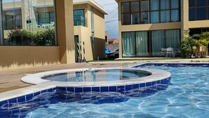 A swimming pool with a tiled edge, a thatched roof structure, and modern buildings with large windows. - Casa milagres Morada do toque (São Miguel dos Milagres)
