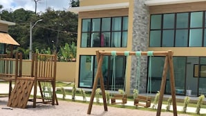 A playground with swings and a slide, a building with large windows, and a sandy area with grass. - Casa milagres Morada do toque (São Miguel dos Milagres)