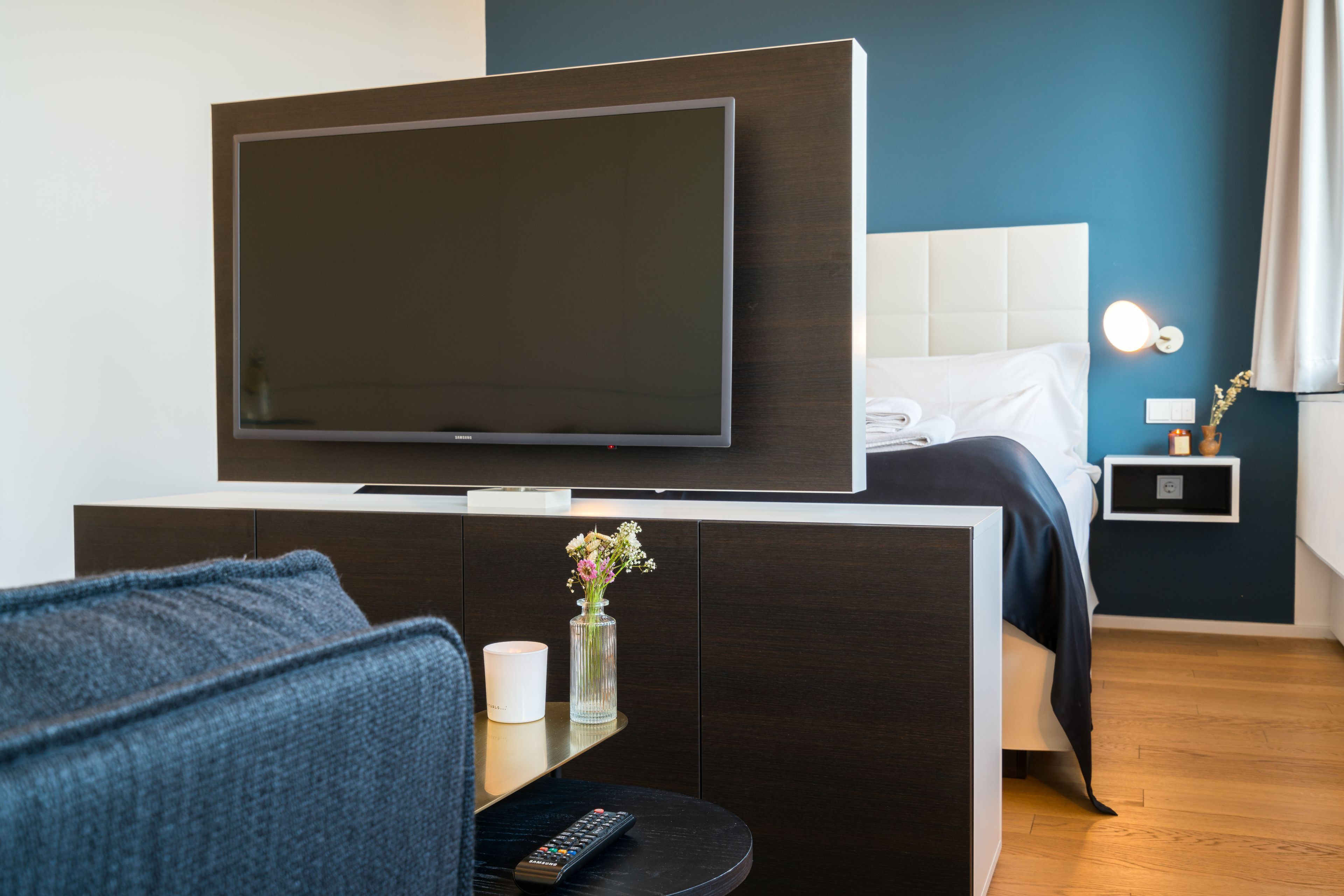 Quarto de hotel com televisão de ecrã plano presa a um móvel de madeira escura, cama com roupa de cama branca, uma pequena mesa de cabeceira com um vaso de flores e um criado-mudo com uma lâmpada.