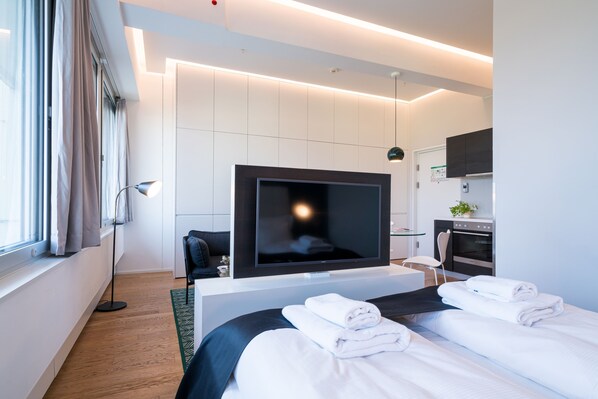 A modern hotel room with a large flat-screen TV, a bed with neatly folded white towels, a small seating area, and a kitchenette.
