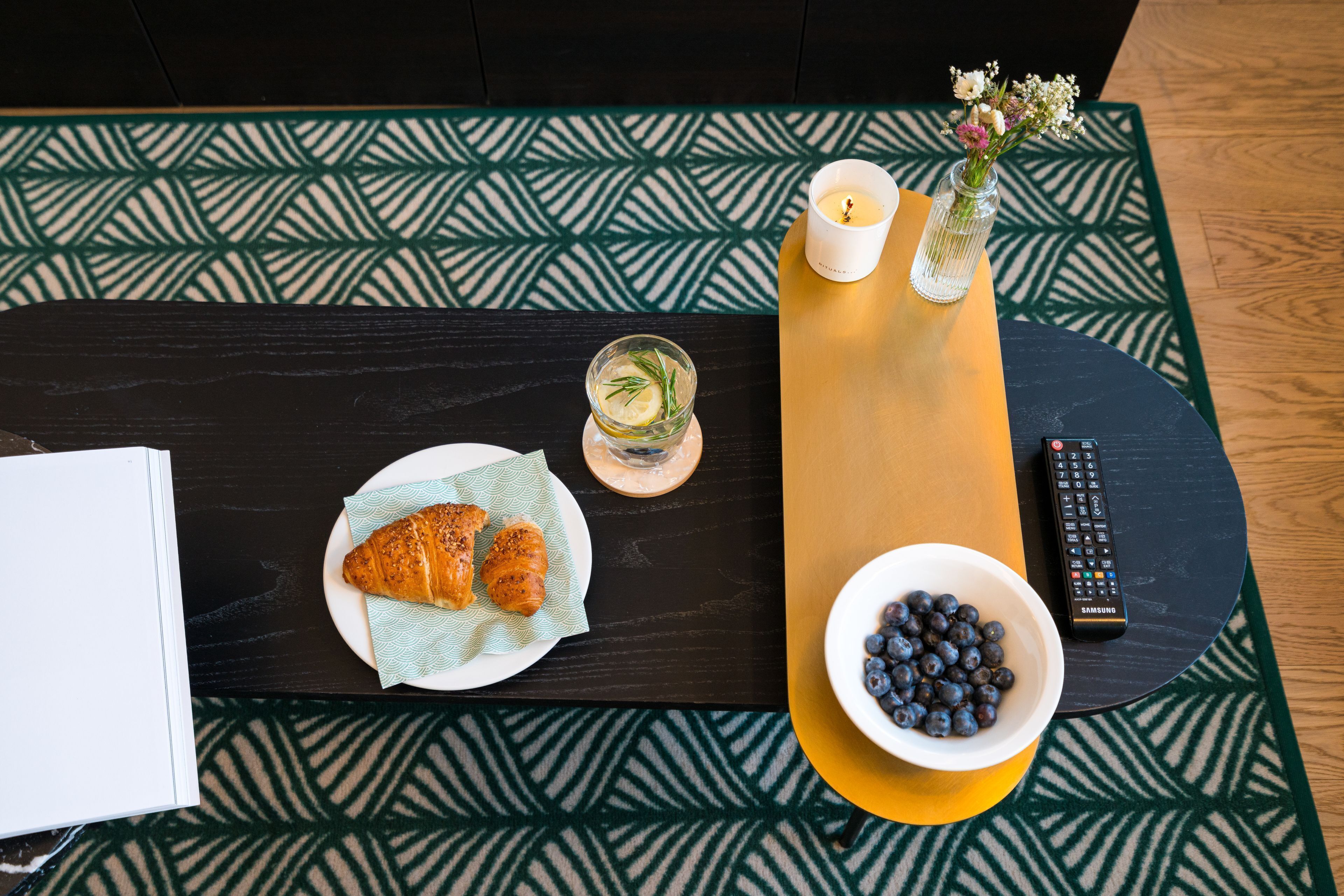 Pöydällä on croissant-leivoksia, kulhollinen mustikoita, kaukosäädin ja kynttilä.