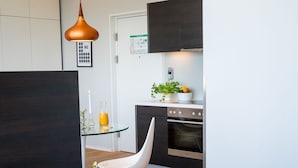 A modern hotel room with a bed, a glass table, a white chair, a kitchen area with cabinets and a stove, and a pendant light. - Vienna Residence, Philips Haus - Euro Plaza #6571 (Vienna)