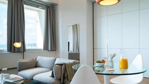 A modern hotel room with a glass dining table, white chairs, a sofa, a small side table with a book and a vase, and a large window with curtains. - Vienna Residence, Philips Haus - Euro Plaza #6571 (Vienna)