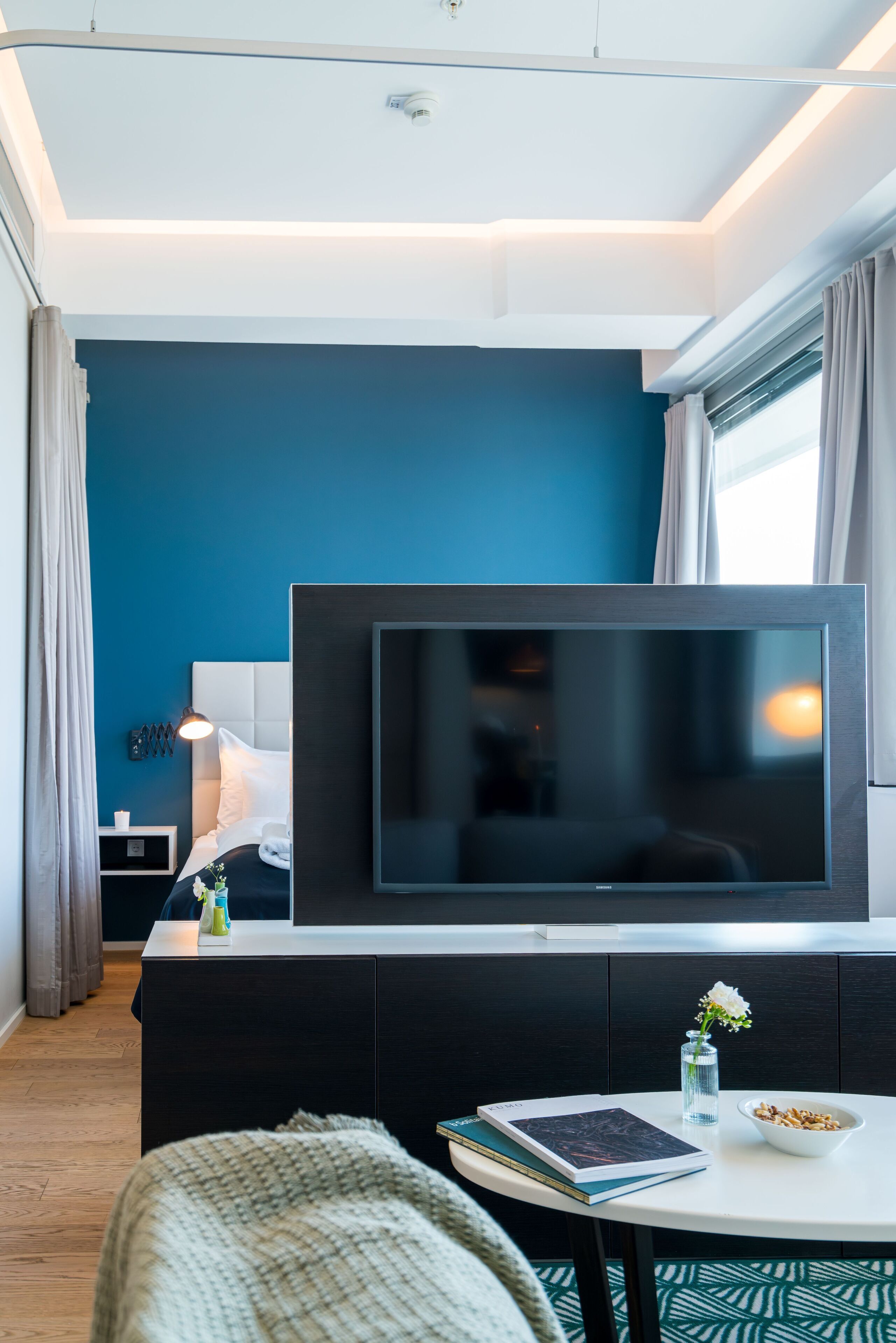 Una habitación de hotel moderna con un televisor de pantalla plana grande, una mesita de noche con un libro y un tazón de cereal, una lámpara de noche pequeña y una ventana con cortinas.