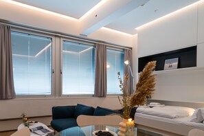 A modern hotel room with a sofa, a glass coffee table, a bed, and large windows with blinds.
