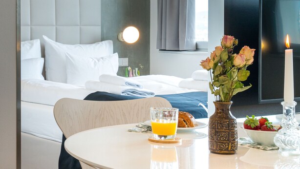 Ein Hotelzimmer mit Bett, Nachttisch mit Blumenvase, Kerze und Frühstücksangeboten.