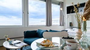 Una habitación de hotel moderna con un ventanal grande, un sofá azul, una mesa de comedor con un plato de comida, una taza de café y un jarrón con plantas secas.