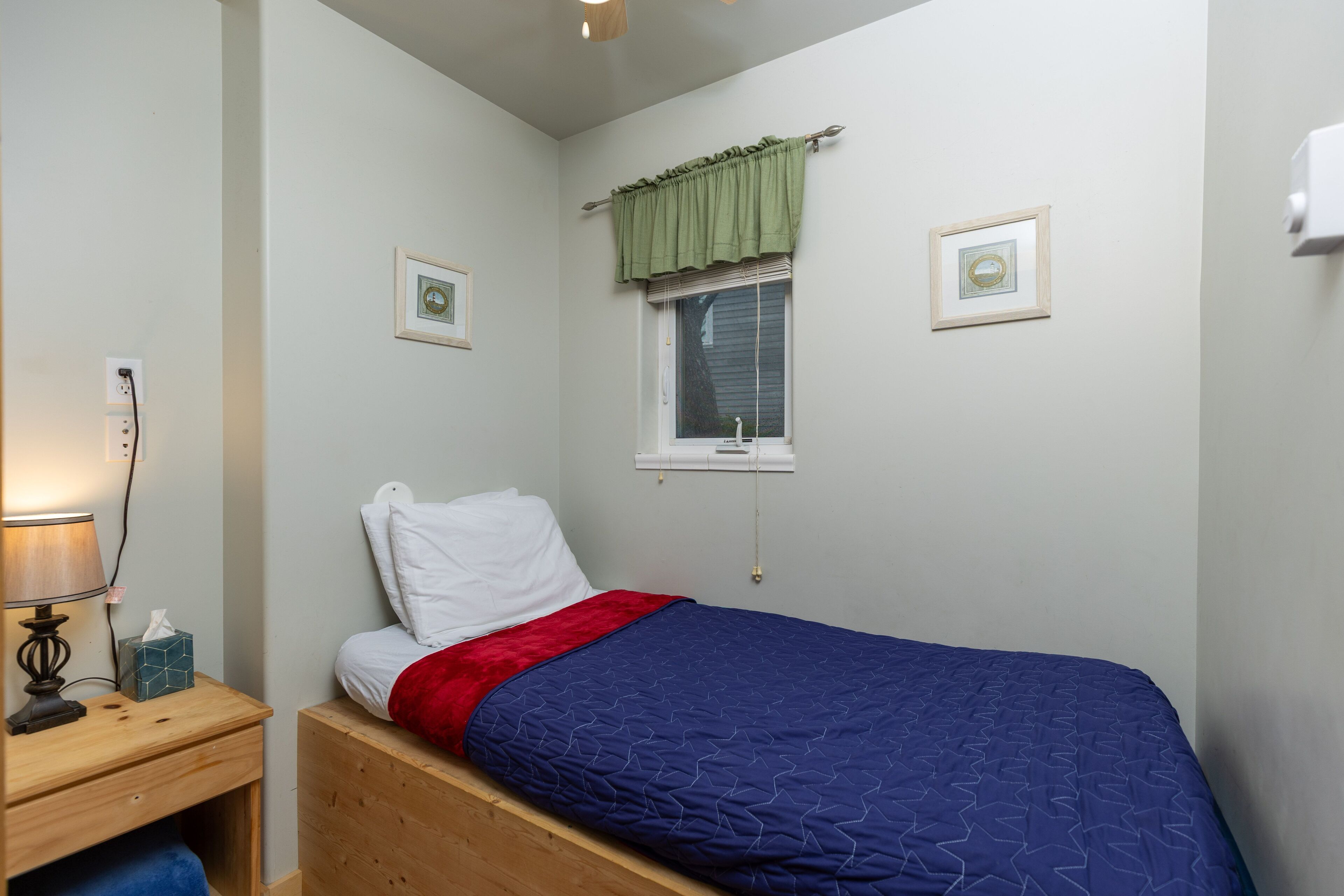 A bedroom with a single bed, a bedside table with a lamp, a window with a curtain, and two framed pictures on the wall.