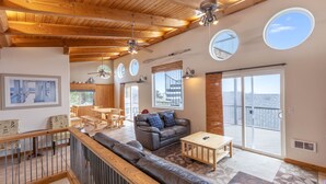 A spacious living area with a wooden ceiling, a leather sofa, a coffee table, and a dining table. There are multiple windows and a sliding glass door leading to an outdoor balcony.