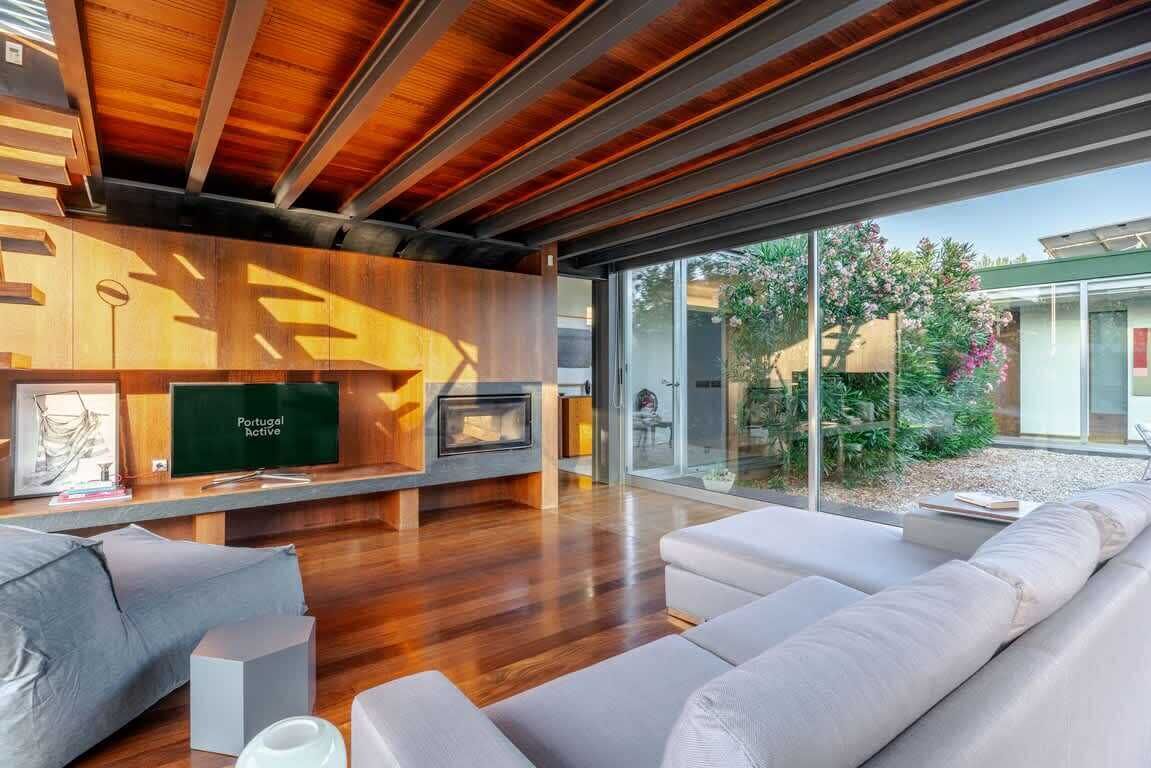 A modern living room with a wooden ceiling, a flat-screen TV, a fireplace, and a large window overlooking a garden.