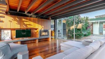 A modern living room with a wooden ceiling, a flat-screen TV, a fireplace, and a large window overlooking a garden.