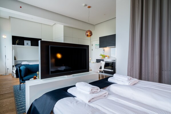 A modern hotel room with a large flat-screen TV, a bed with neatly folded towels, a desk with a chair, and a kitchenette in the background.