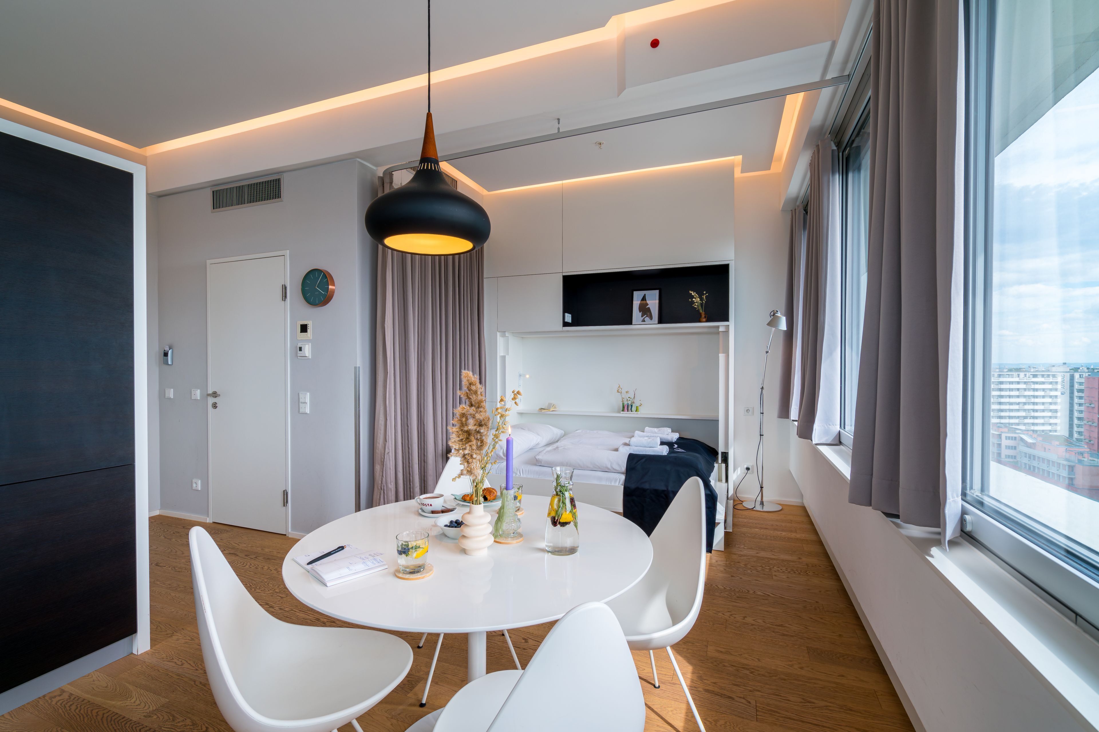 Moderni hotellihuone, jossa on pyöreä ruokapöytä, valkoiset tuolit, riippuvalaisin ja suuri ikkuna, jossa on verhot.
