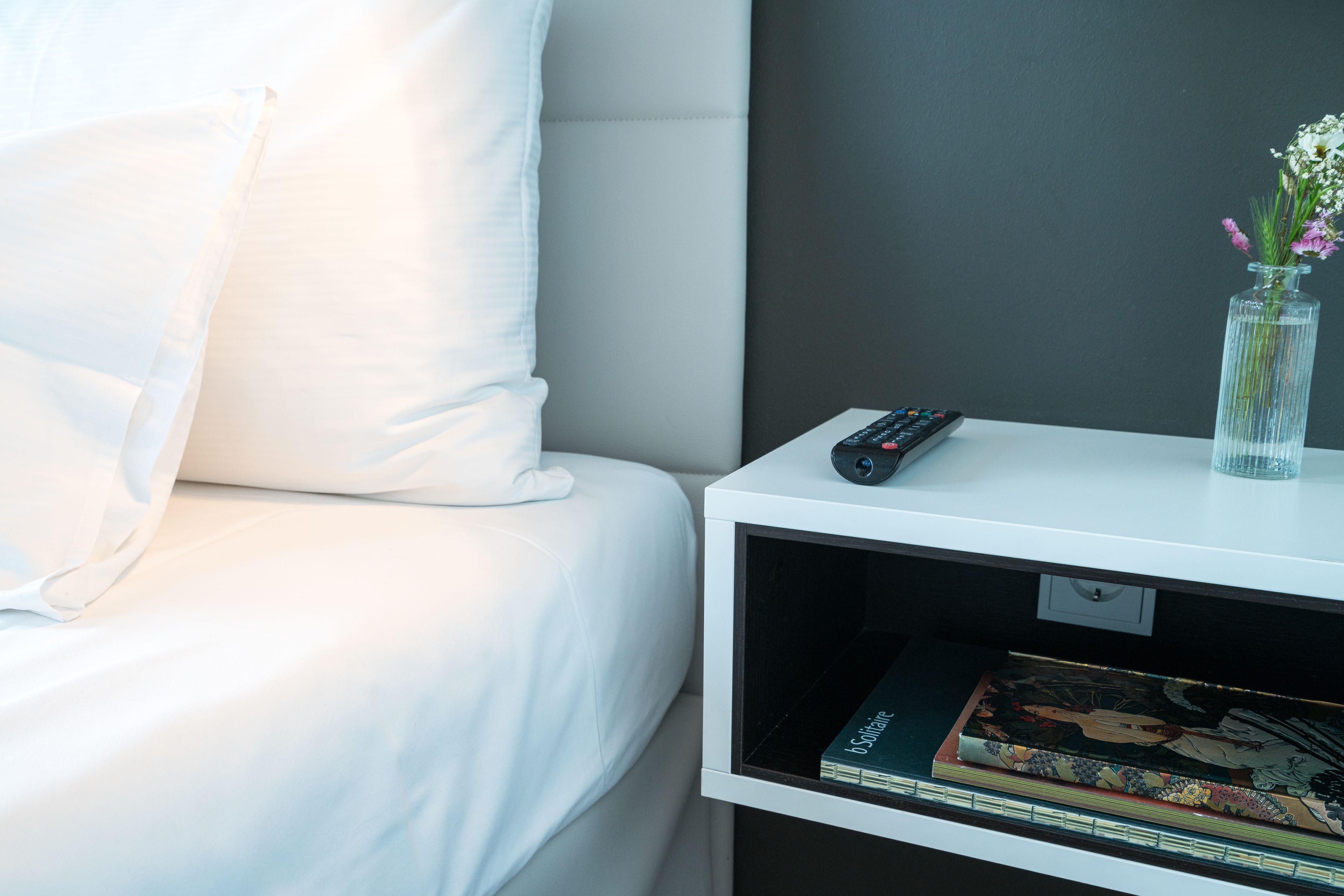 A neatly made bed with white linens, a white nightstand with a remote control, a vase with flowers, and a book on the nightstand.