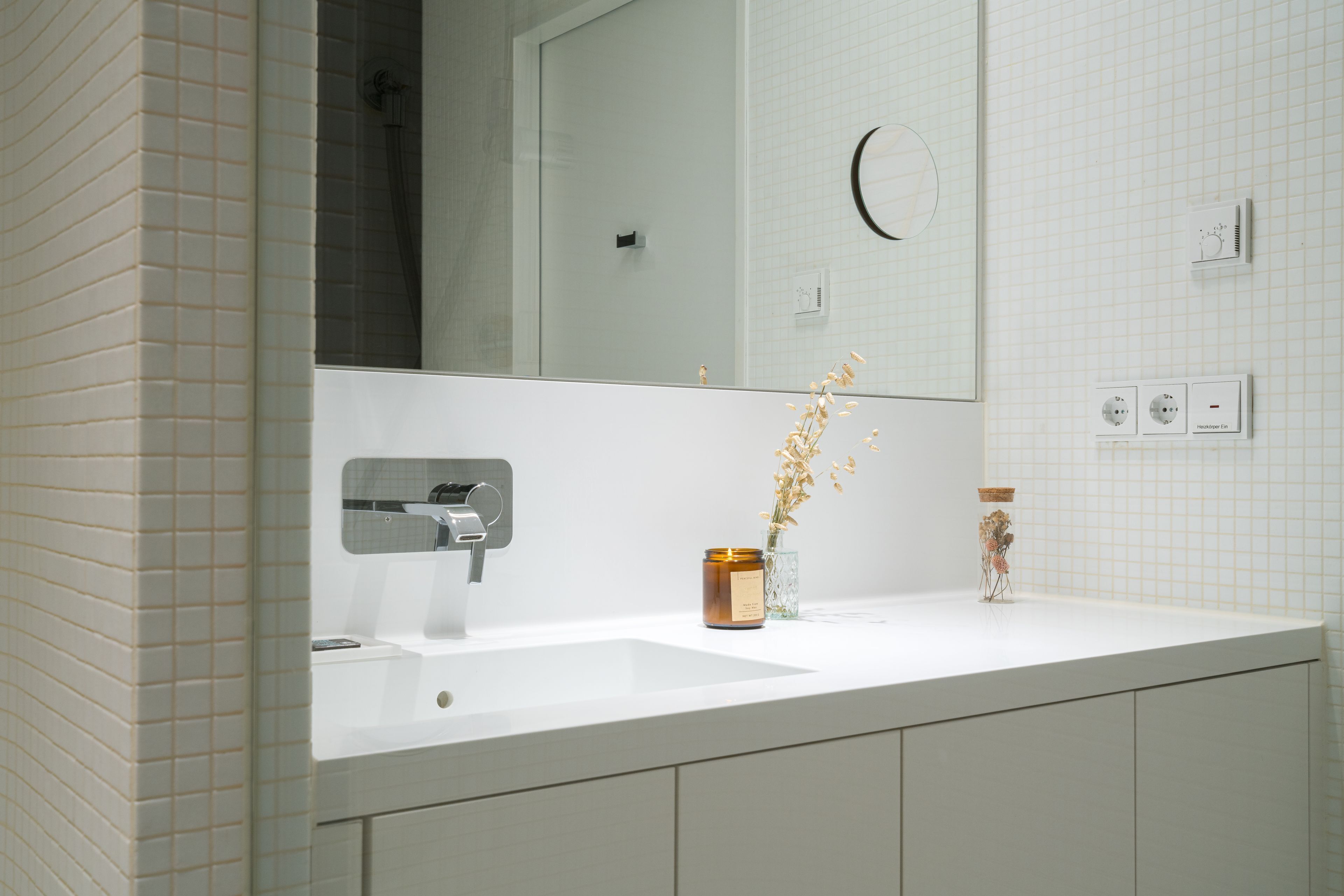 Ein Badezimmer mit einem weißen Waschbecken, einem Spiegel und einer Dusche. Auf dem Waschbecken liegen zwei kleine Dekorationen.