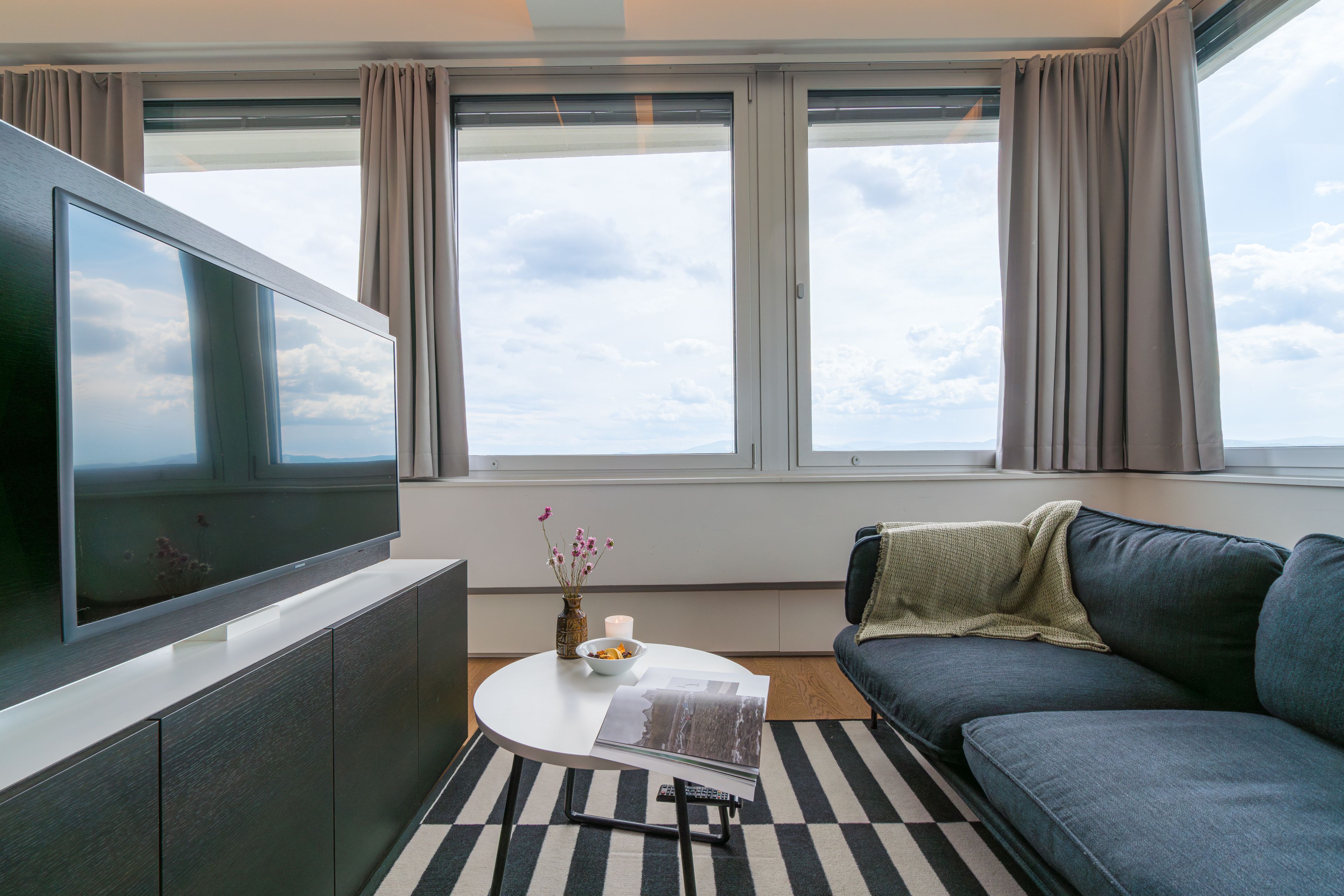 A modern living room with a flat-screen TV, a sofa, a coffee table, and a view of the sky through large windows.