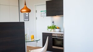 A modern hotel room with a bed, a glass table, a white chair, a kitchen area with cabinets and a stove, and a pendant light. - Vienna Residence, Philips Haus - Euro Plaza #6526 (Vienna)