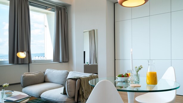 A modern hotel room with a glass dining table, white chairs, a sofa, a small side table with a book and a vase, and a large window with curtains. - Vienna Residence, Philips Haus - Euro Plaza #6526 (Vienna)