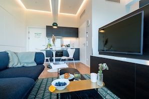 A modern living room with a sofa, a dining table with a bowl of fruit, a TV, and a kitchen in the background.