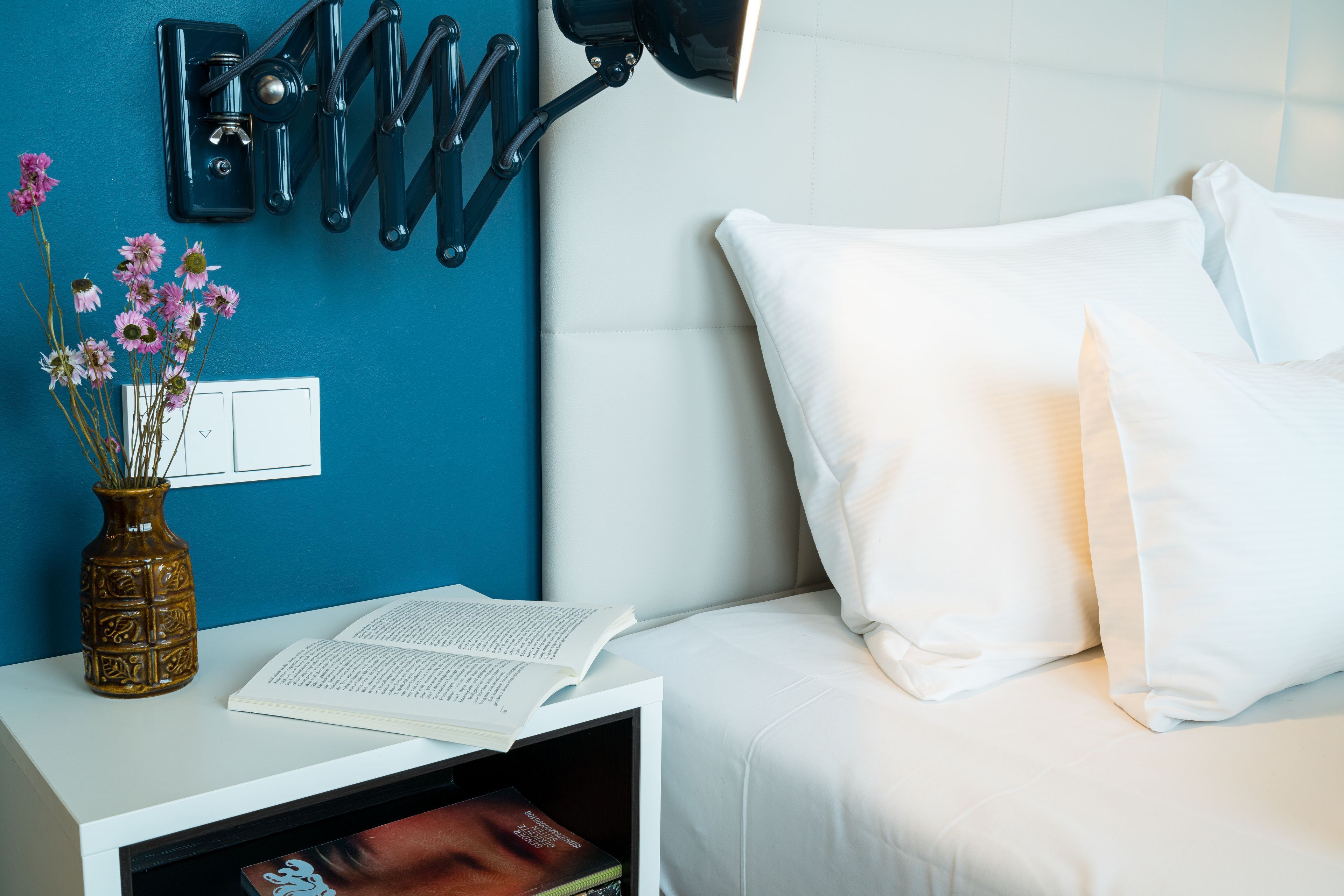 A neatly arranged hotel room with a white bedside table, a book, a vase with flowers, and a wall-mounted lamp.