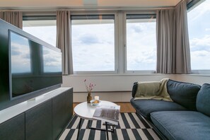 En moderne stue med et fladskærms-tv, en sofa, et sofabord og udsigt til himlen gennem store vinduer.