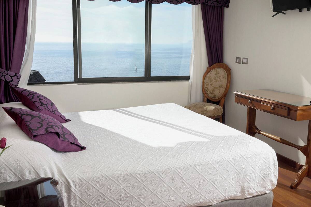 A bedroom with a large bed, a wooden nightstand, a chair, and a view of the sea through a window.
