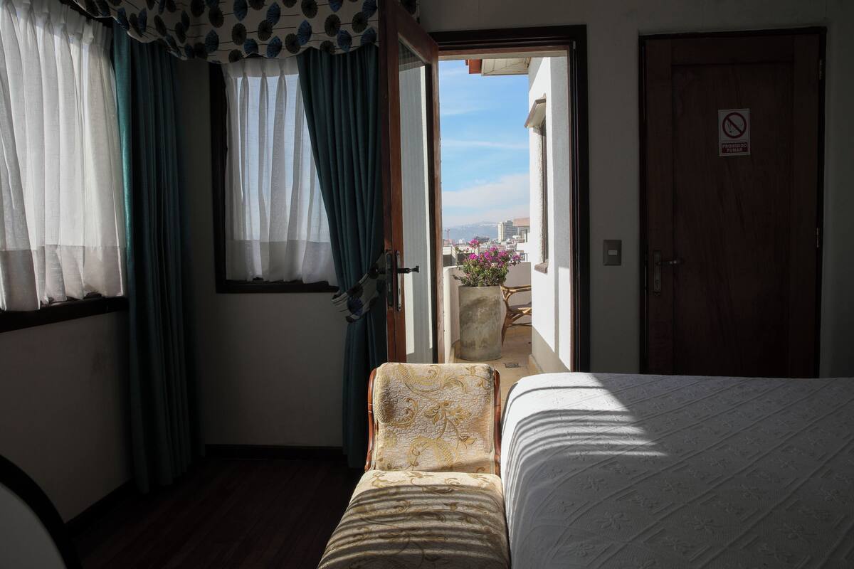 A hotel room with a bed, a chair, a balcony with a view, and a door with a no smoking sign.