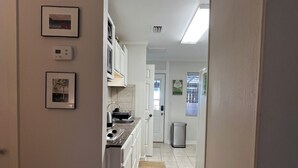 A narrow hallway with a kitchen area on the left, featuring white cabinetry, a stainless steel sink, and a small rug leading to a door.