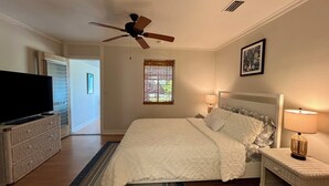A bedroom with a bed, a television, a ceiling fan, a window with blinds, and a nightstand with a lamp.