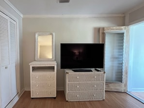 Une pièce avec un téléviseur posé sur un meuble, un miroir et une fenêtre avec des stores.