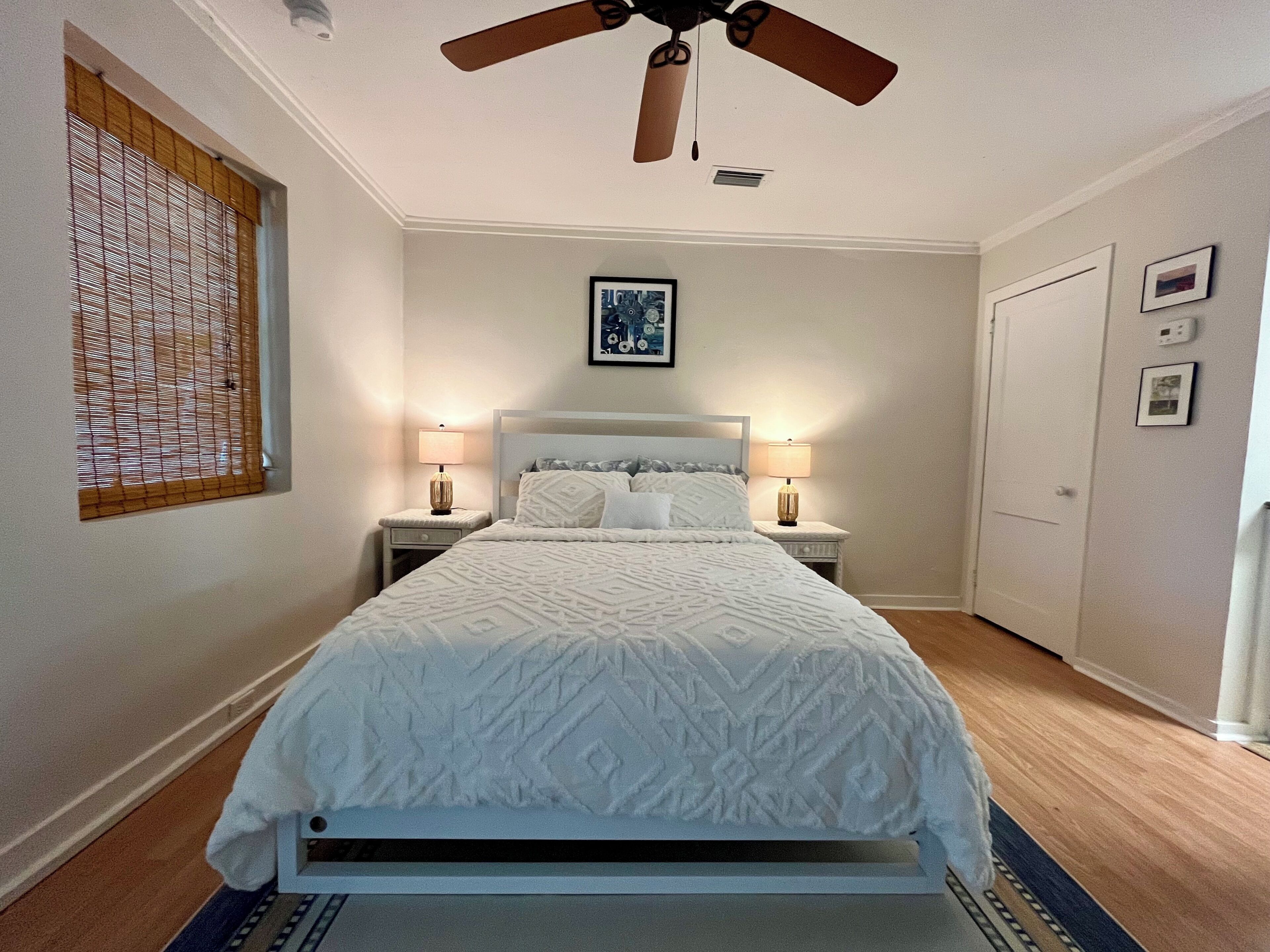 A bedroom with a bed, two bedside tables with lamps, a ceiling fan, a window with blinds, and a door.