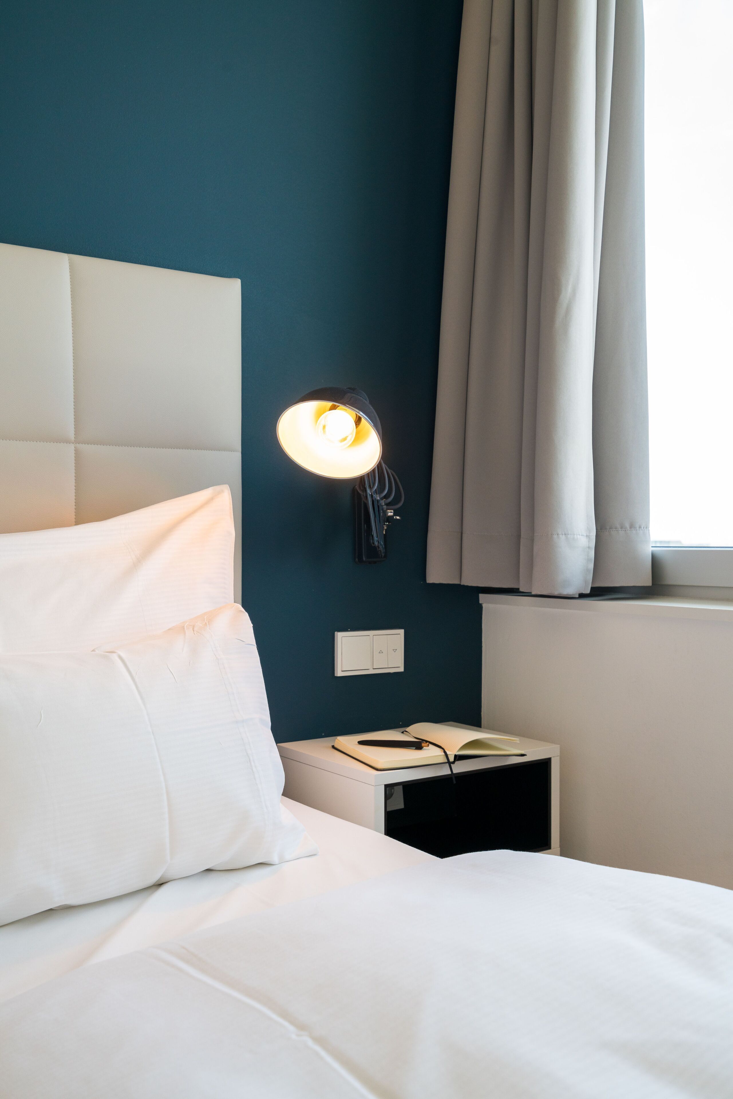 Ein ordentlich bezogenes Bett mit weißen Leinen, eine Nachttischlampe und ein Nachttisch mit einem Buch und einem Telefon.
