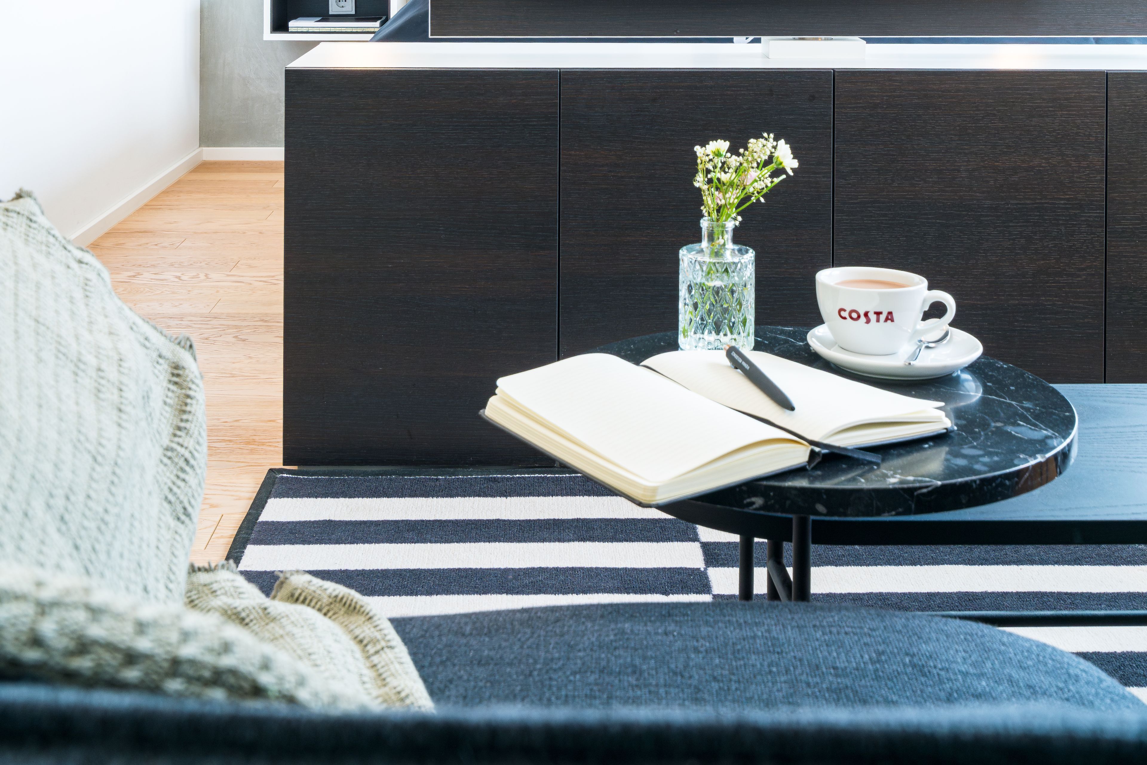Ein modernes Wohnzimmer mit einem dunklen Holzschrank, einem runden schwarzen Tisch mit einem Buch und einer Tasse mit dem Schriftzug „COSTA“ sowie einem Streifen Teppich.