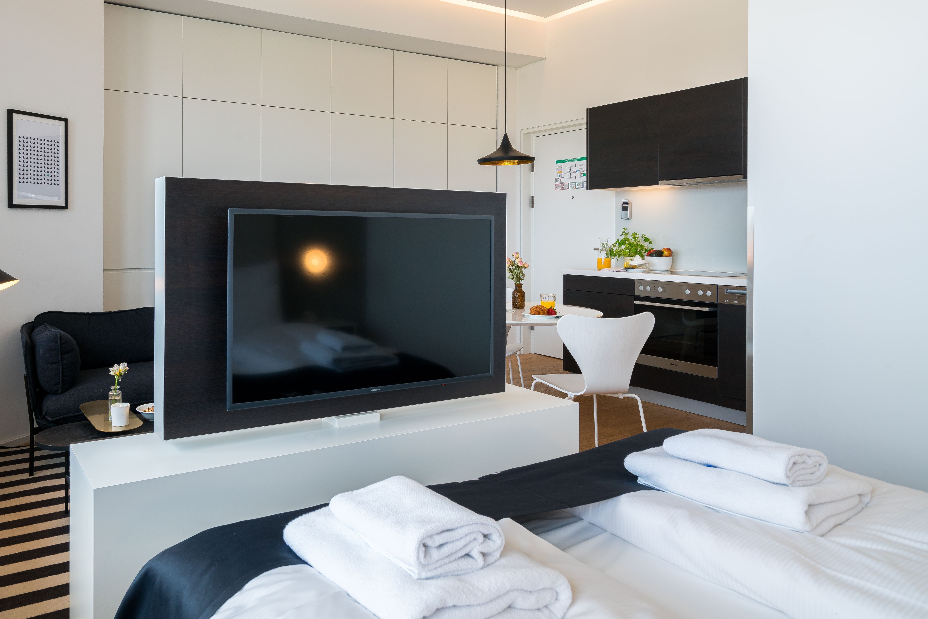 Ein modernes Hotelzimmer mit einem großen Flachbildfernseher, einem Bett mit weißer Bettwäsche, einer kleinen Sitzecke mit Sofa und Couchtisch sowie einer Küchenzeile mit Herd und Mikrowelle.
