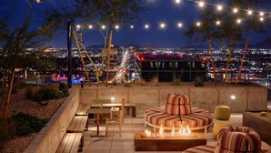 An outdoor seating area with a fire feature, wooden furniture, and string lights against a cityscape view. - Sentral Sol Modern (Phoenix)