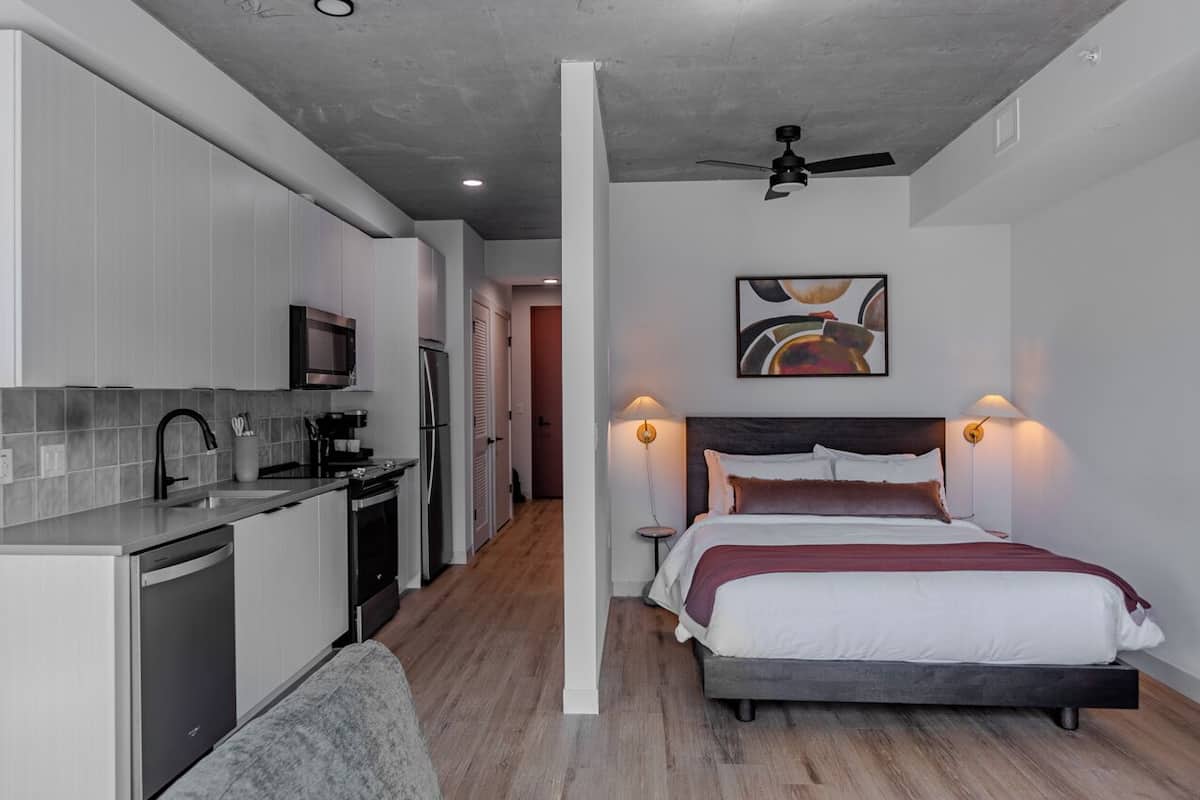 A modern hotel room with a kitchen, a bed with a headboard, and a ceiling fan.