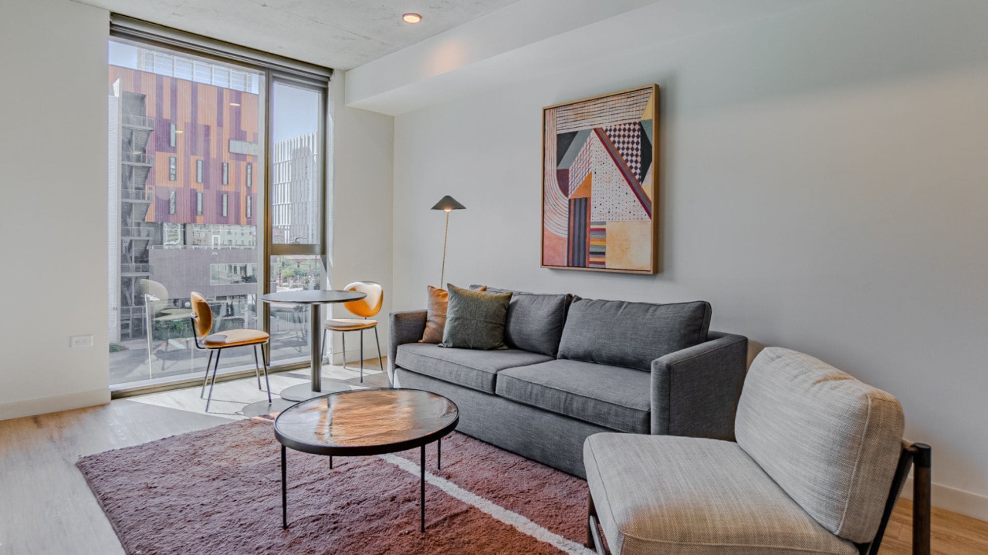 Un salon moderne avec un canapé gris, une table basse ronde et deux fauteuils. Une grande fenêtre offre une vue sur le paysage urbain.