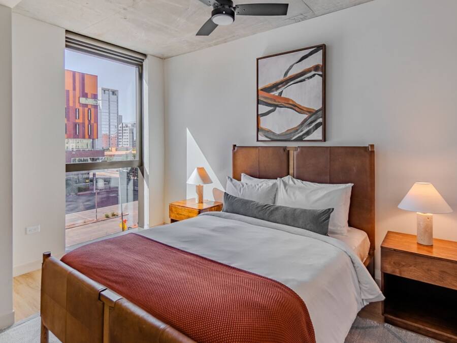 A hotel room with a bed, a nightstand, a lamp, a ceiling fan, a window with a city view, and a framed artwork on the wall.
