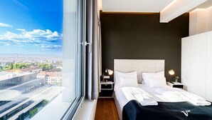 Et moderne hotelværelse med et stort vindue, en seng med hvide sengetøj, et natbord med en lampe og udsigt over bybilledet.