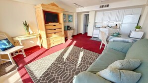 A living room with a sofa, a wooden cabinet, a television, and a kitchen area with cabinets and appliances.